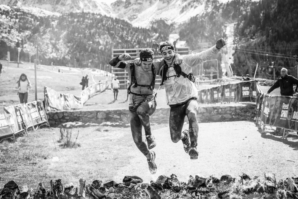 two men with arms round each other jumping over an obstacle